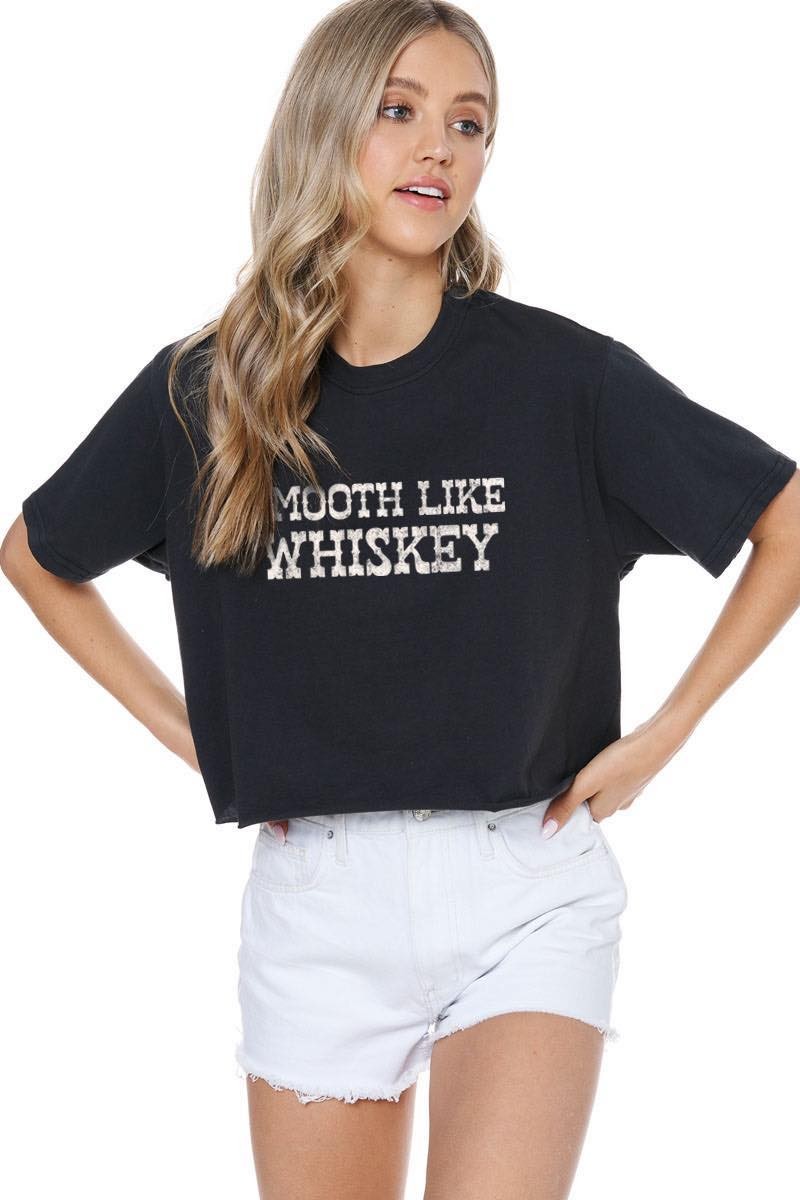 A woman modeling a black cropped t-shirt with 'SMOOTH LIKE WHISKEY' printed in distressed white letters across the chest, paired with white denim shorts against a plain white background.