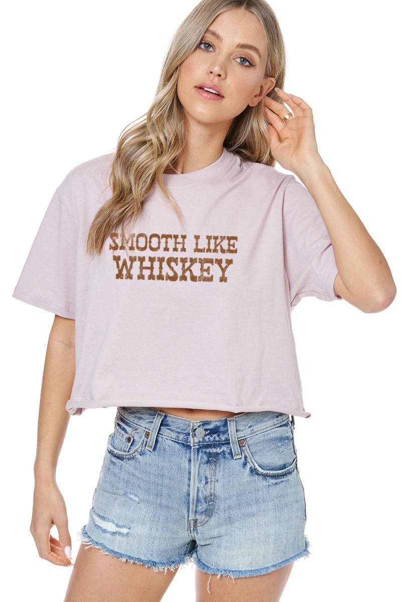 A woman modeling a dusty rose cropped t-shirt with 'SMOOTH LIKE WHISKEY' printed in brown across the chest, paired with light-wash denim shorts against a white background.