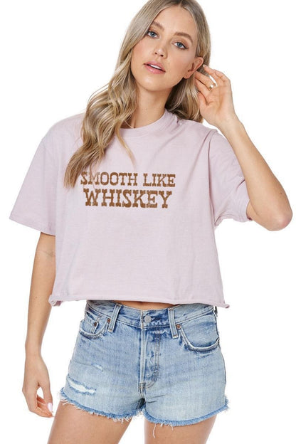 A woman modeling a dusty rose cropped t-shirt with 'SMOOTH LIKE WHISKEY' printed in brown across the chest, paired with light-wash denim shorts against a white background.