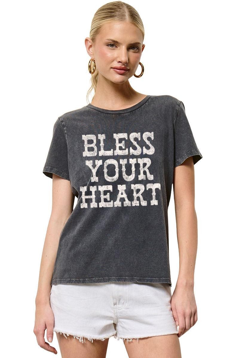 A woman modeling a dark gray acid-wash graphic t-shirt with the text 'BLESS YOUR HEART' in distressed white letters, paired with white denim shorts against a plain white background.