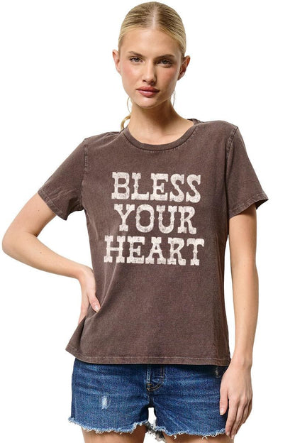 A woman modeling a brown acid-wash t-shirt with 'BLESS YOUR HEART' printed in distressed white block letters, paired with blue denim shorts against a white background.