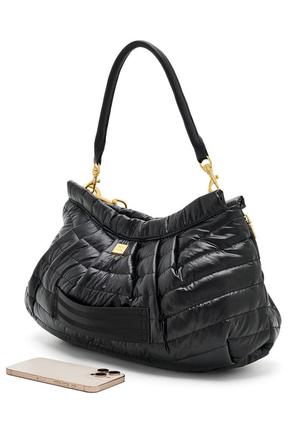 A black quilted hold me close clutch puffer handbag with gold-tone hardware and a black shoulder strap, shown on a white background with a smartphone placed in front for scale.