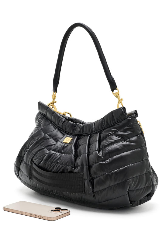 A black quilted hold me close clutch puffer handbag with gold-tone hardware and a black shoulder strap, shown on a white background with a smartphone placed in front for scale.