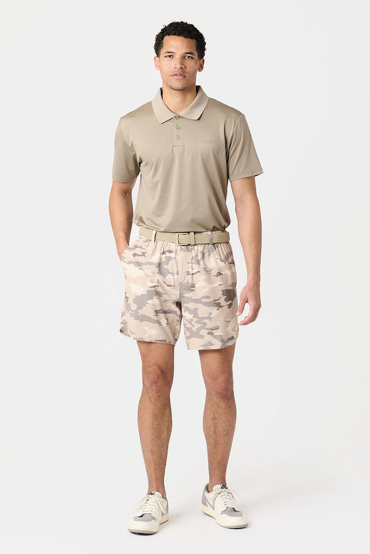 A man modeling a beige short-sleeve polo shirt and matching beige river rock camouflage shorts with a light tan belt, paired with white sneakers, against a plain light gray studio background.