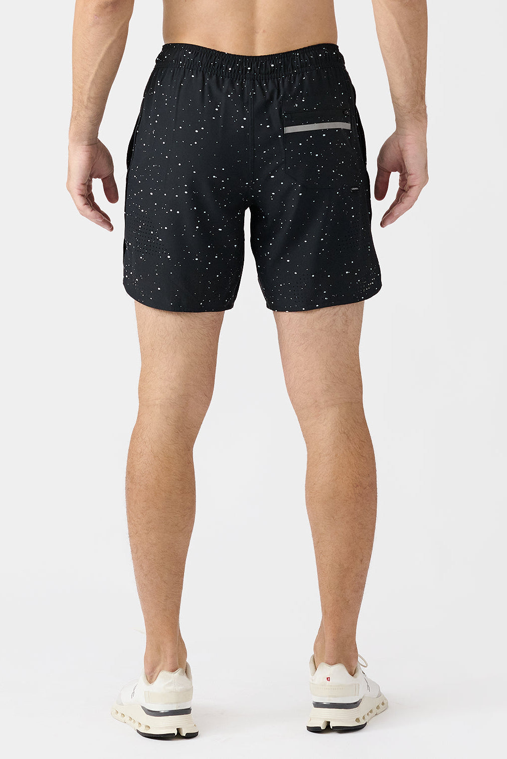 A man viewed from the back wearing black athletic shorts with a white splatter pattern and white sneakers, set against a plain light gray studio background.