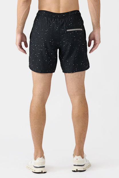 A man viewed from the back wearing black athletic shorts with a white splatter pattern and white sneakers, set against a plain light gray studio background.