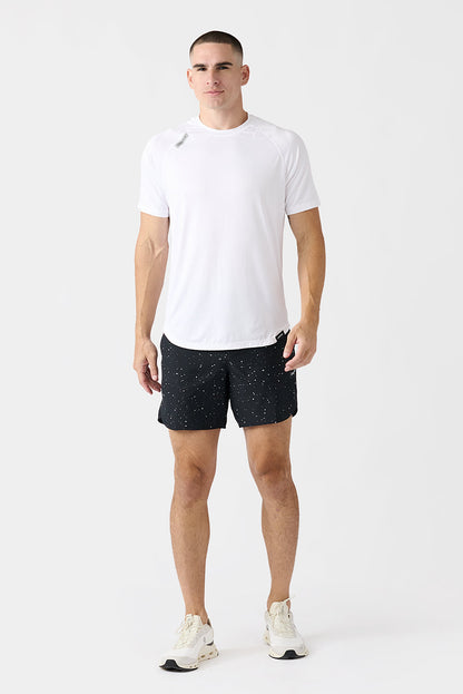 A man modeling a white short-sleeve athletic t-shirt and black & white splattered Luka running shorts with white sneakers, standing against a plain light gray studio background.