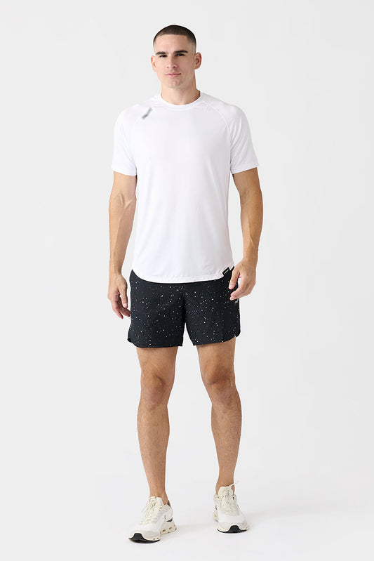 A man modeling a white short-sleeve athletic t-shirt and black & white splattered Luka running shorts with white sneakers, standing against a plain light gray studio background.