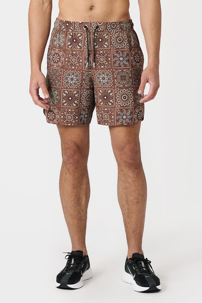 A man modeling a pair of copper brown paisley tile patterned shorts with a drawstring waist, paired with black athletic sneakers, against a plain light gray studio background.