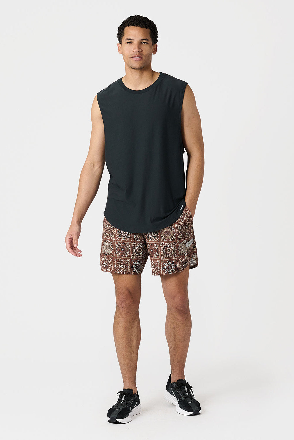 Man wearing a black sleeveless shirt and paisley tile copper patterned shorts on a white background