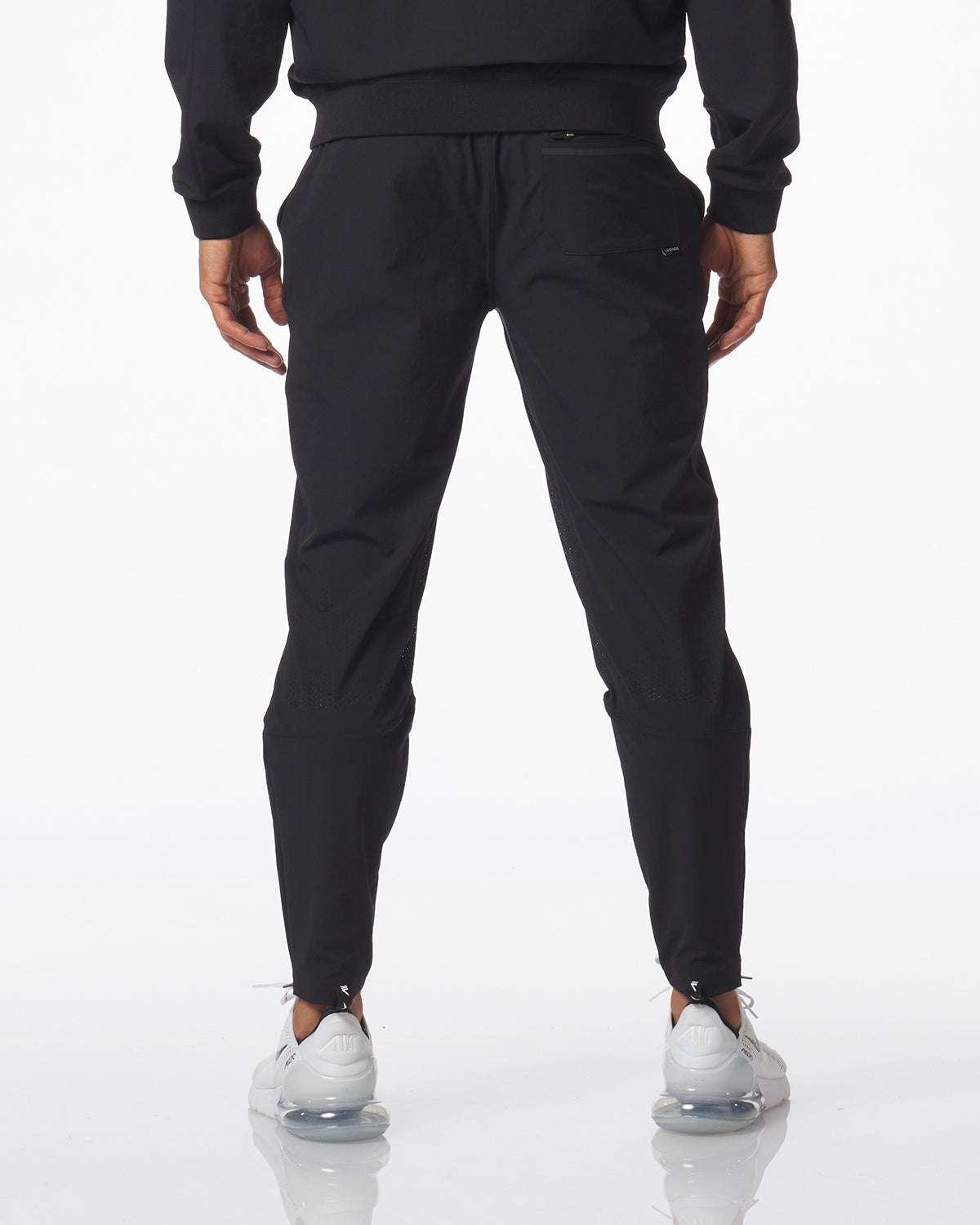 Rear view of a person wearing black jogger-style carson pants with a drawstring waist, paired with a black sweatshirt and white sneakers, against a clean white studio background.