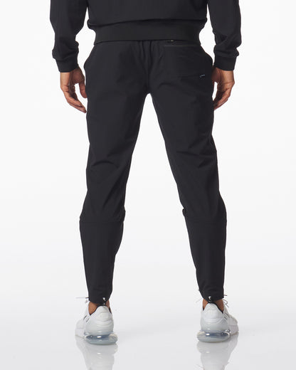 Rear view of a person wearing black jogger-style carson pants with a drawstring waist, paired with a black sweatshirt and white sneakers, against a clean white studio background.