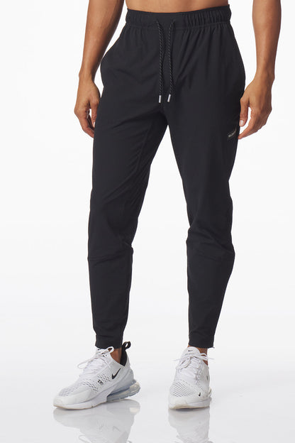 A person modeling black jogger-style carson athletic pants with a drawstring waist and elastic cuffs, paired with white sneakers, against a clean white studio background.