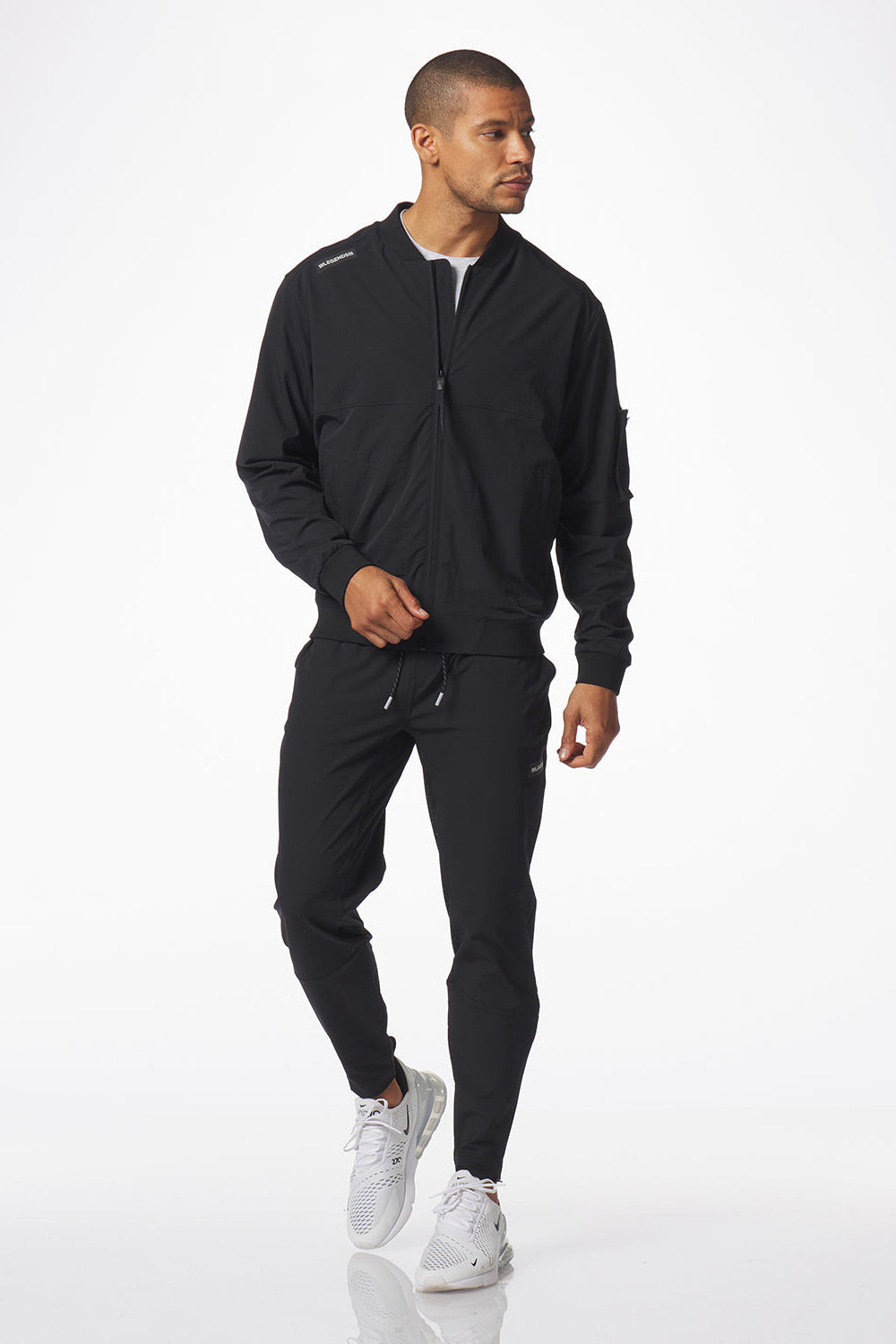 A man modeling a black zip-up jacket and matching black carson  jogger pants with white sneakers, photographed against a clean white studio background.