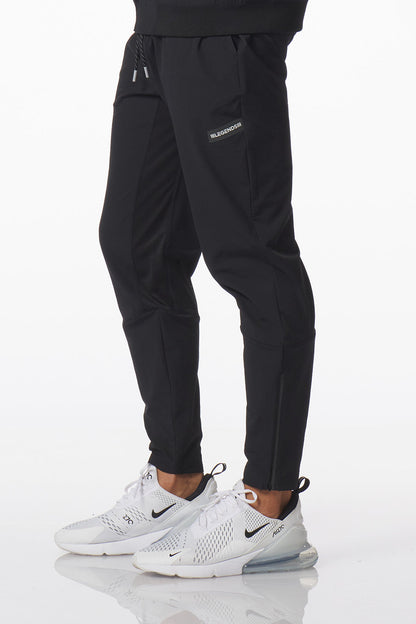 Black Carson athletic jogger pants with side cargo pockets and a small white logo on the upper left thigh, shown on a model wearing white Nike sneakers against a white studio background.