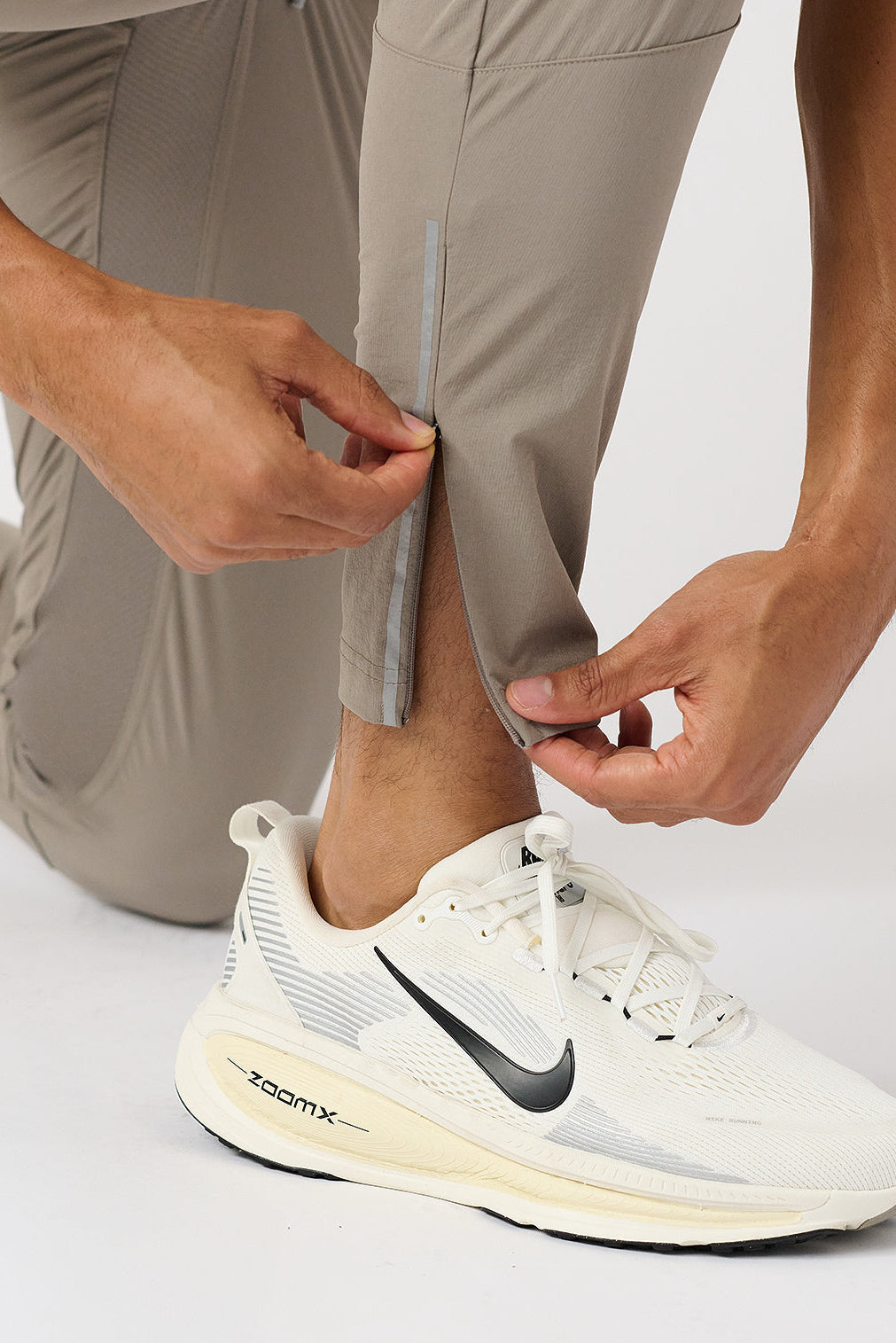 Person wearing taupe pants and a white running shoe with a black Nike logo on a light gray background