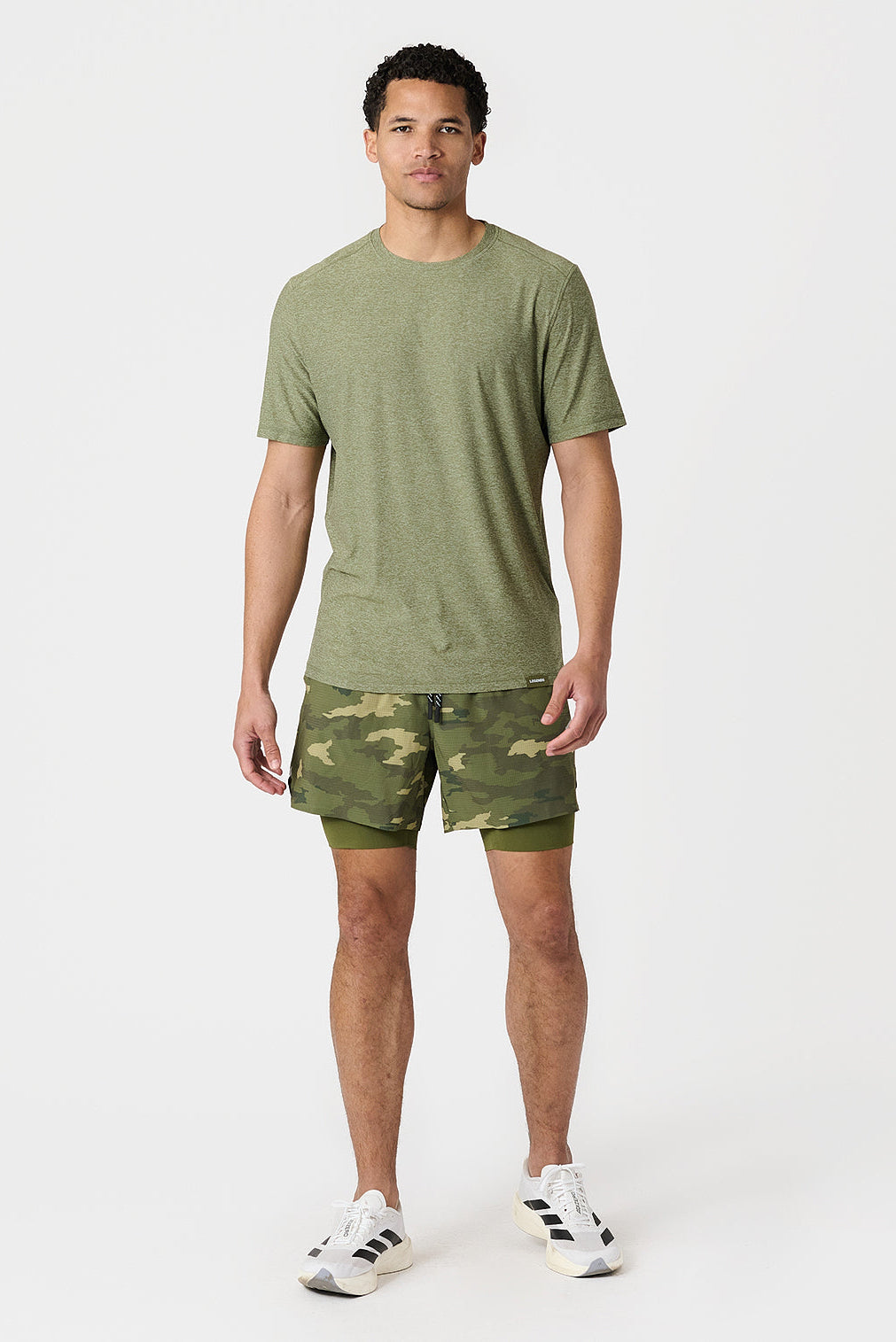 A man modeling a cypress heather crewneck t-shirt and matching camouflage-patterned athletic shorts with a built-in compression layer, paired with white sneakers, against a plain light gray studio background.