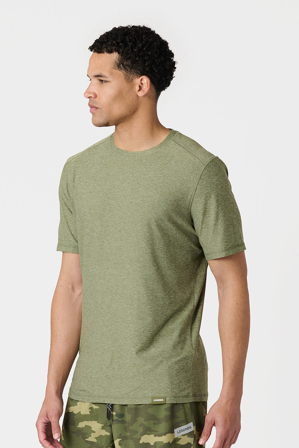 A man wearing a cypress heather crewneck t-shirt and camouflage shorts, shown from a side profile against a light gray studio background.