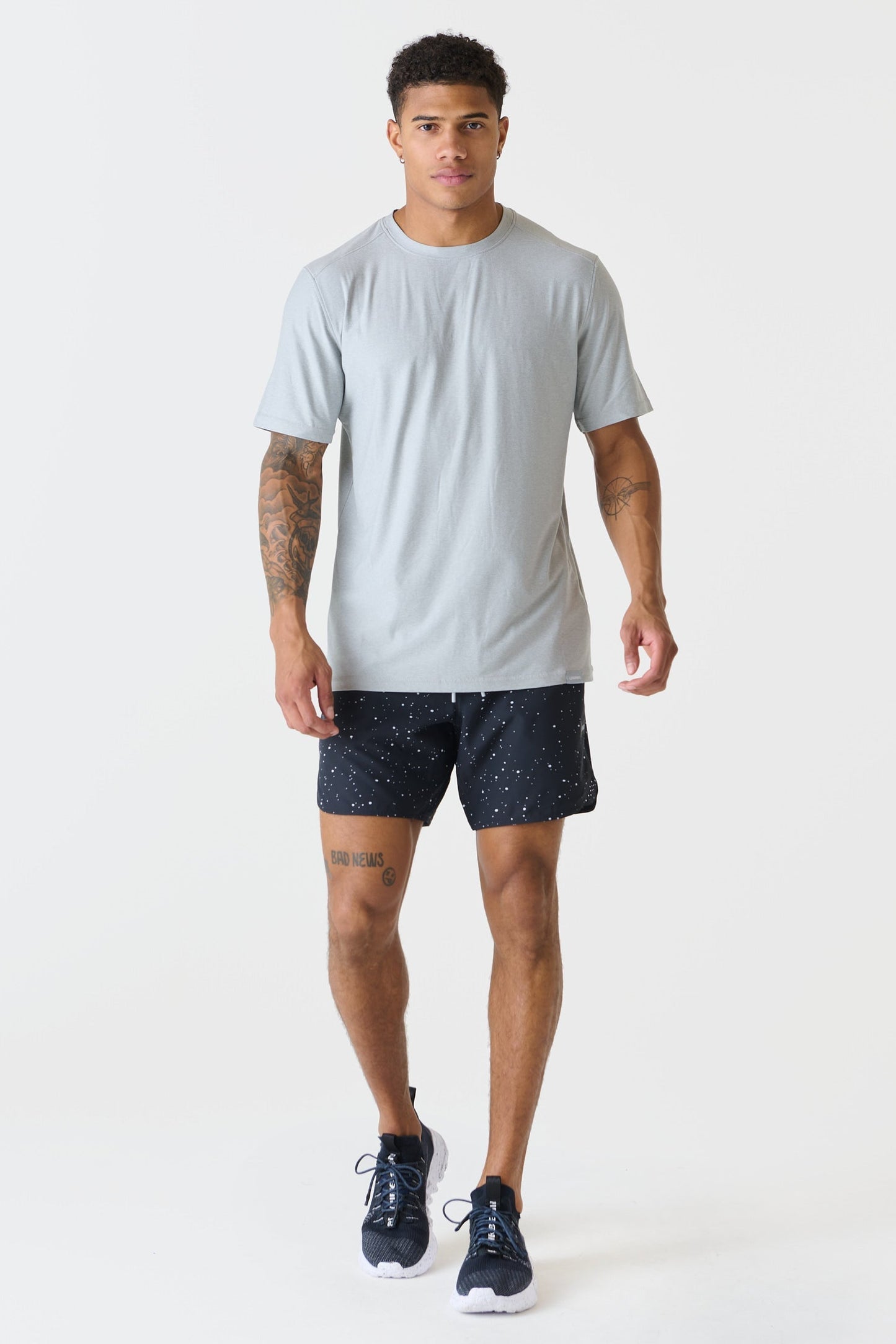A man modeling a heather gray crewneck t-shirt and dark navy patterned athletic shorts with navy running shoes, shot against a solid white studio background.