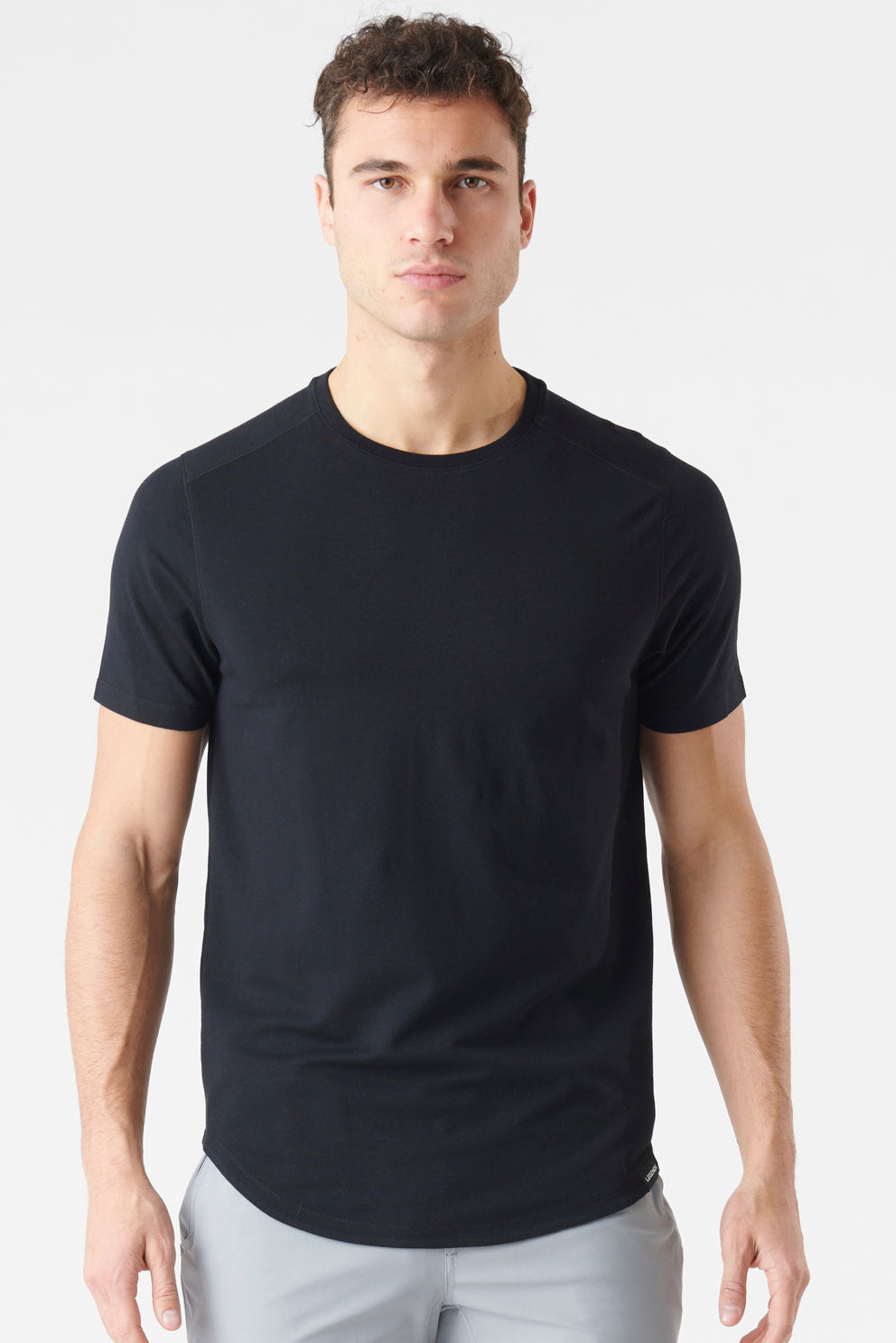 A man wearing a plain black short-sleeve crewneck t-shirt and light gray trousers, standing against a light gray studio background.