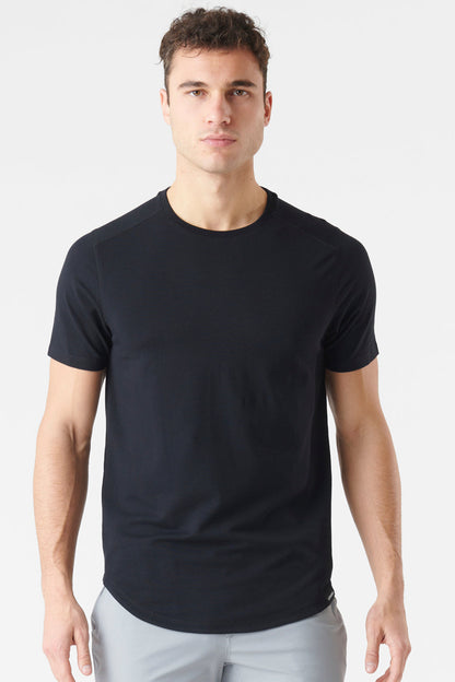 A man wearing a plain black short-sleeve crewneck t-shirt and light gray trousers, standing against a light gray studio background.