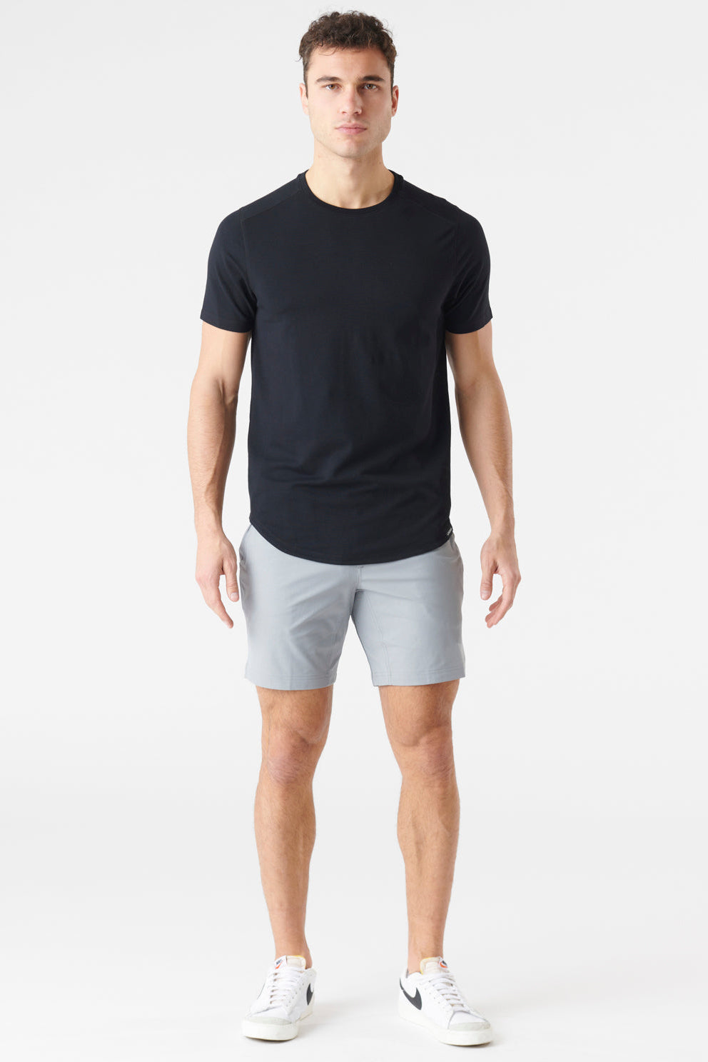 A man modeling a black short-sleeve crewneck t-shirt with a curved hem, light gray shorts, and white sneakers against a plain white studio background.