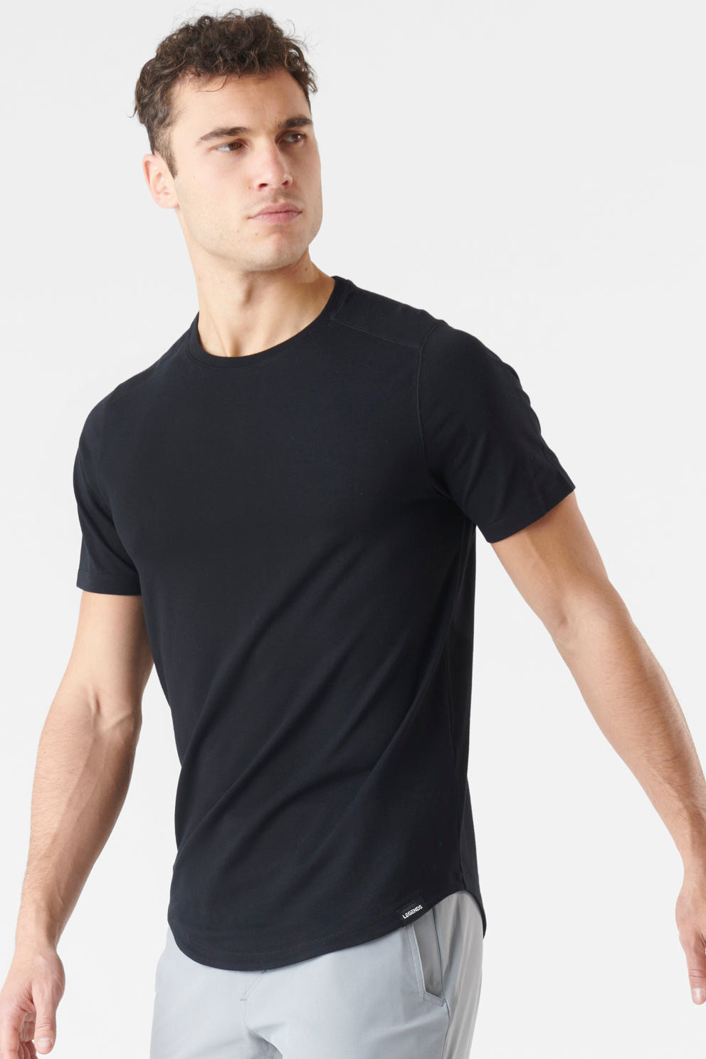 A man wearing a black short-sleeve crewneck athletic t-shirt and light gray pants, photographed against a clean white studio background.