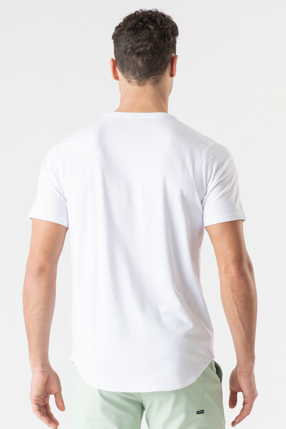 A man viewed from behind wearing a plain white short-sleeve t-shirt and light green shorts against a clean white studio background.