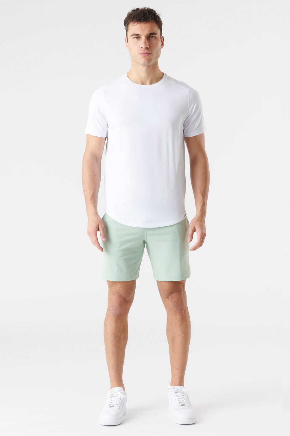 A man modeling a white short-sleeve crewneck t-shirt and light sage green shorts against a plain white studio background.