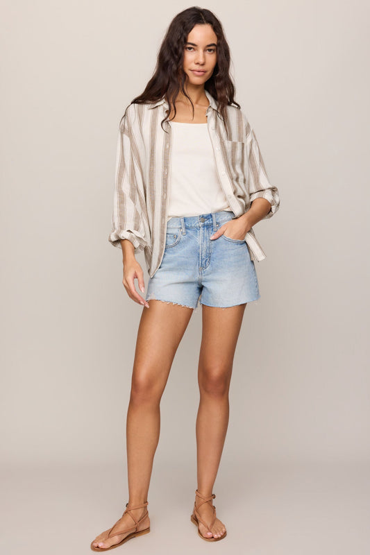 Woman wearing a light toffee striped shirt, white top, and denim shorts on a plain background