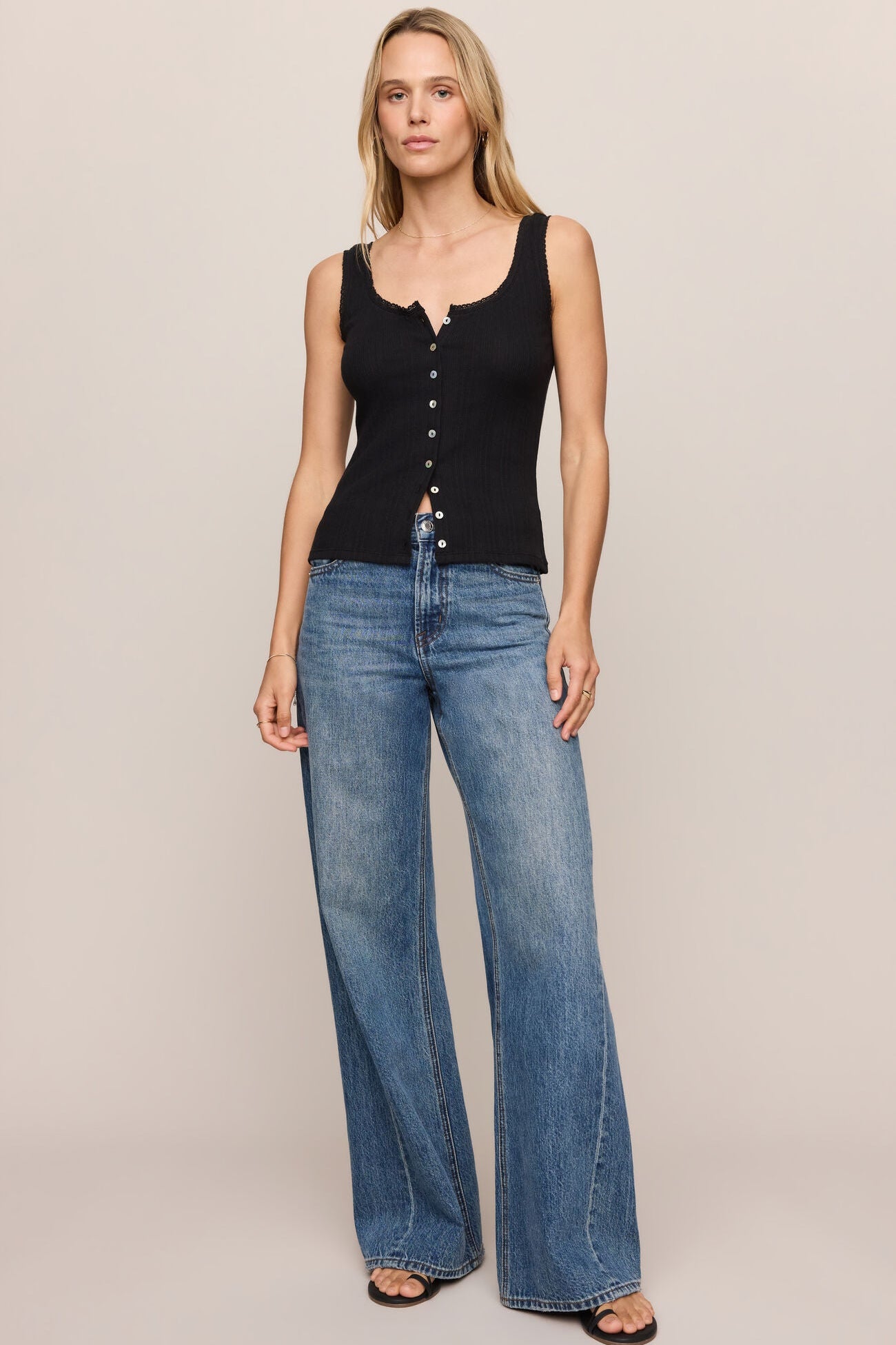 A woman modeling a black sleeveless button-front tank top and wide-leg blue jeans against a plain beige studio background.