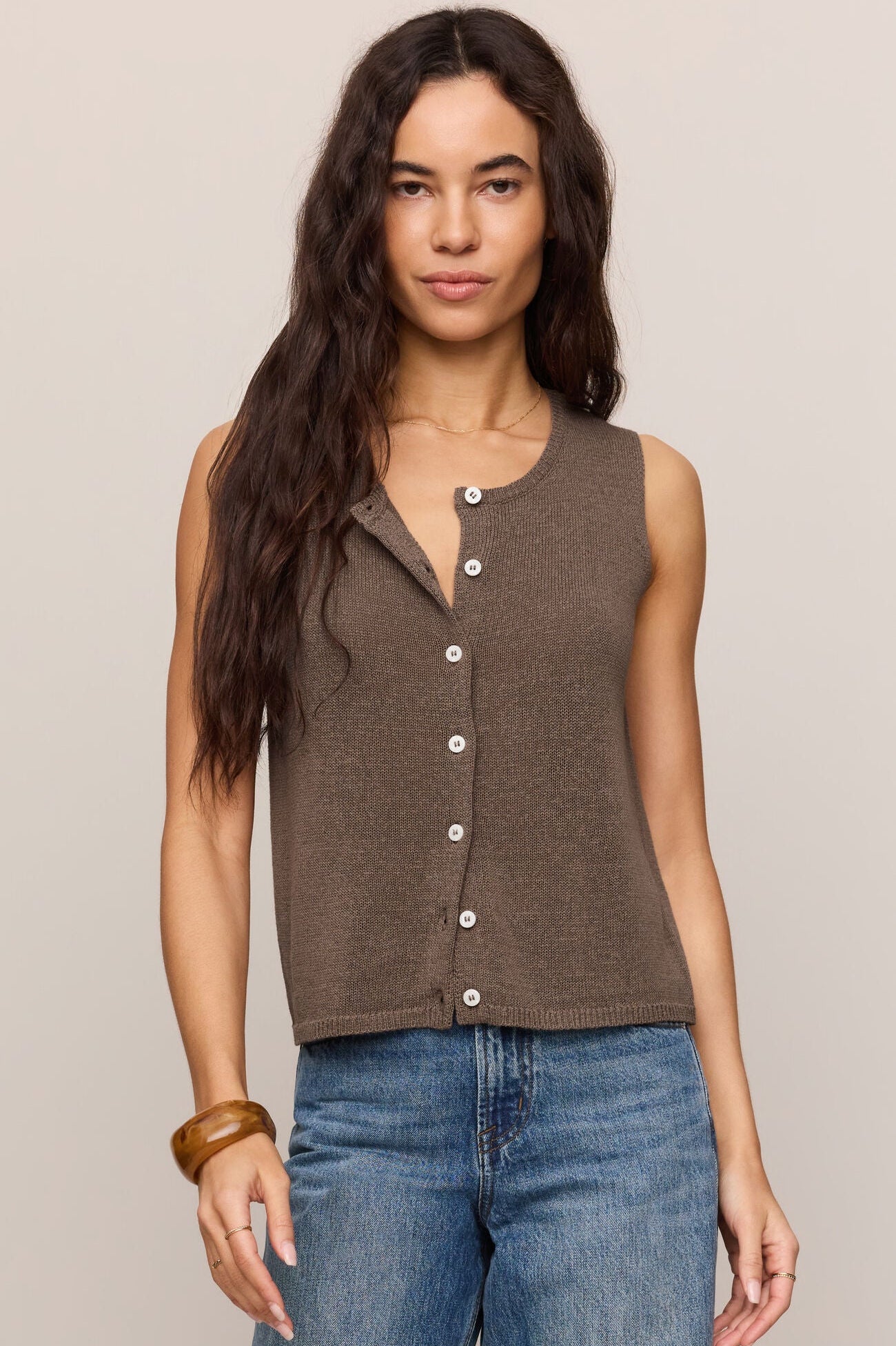 Woman wearing a pebble coast brown sleeveless button-up vest style shirt and blue jeans against a beige background