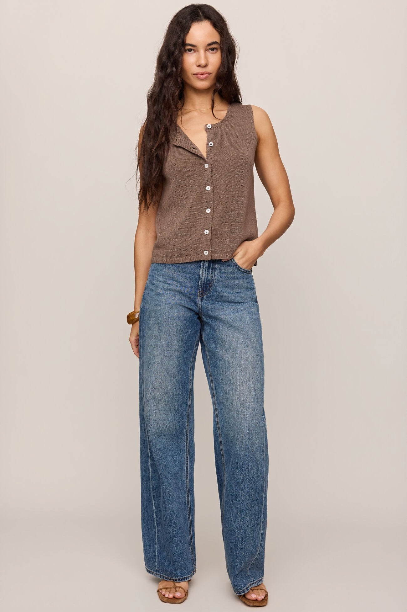 Woman wearing a pebble coast sleeveless button up vest top and blue jeans on a beige background