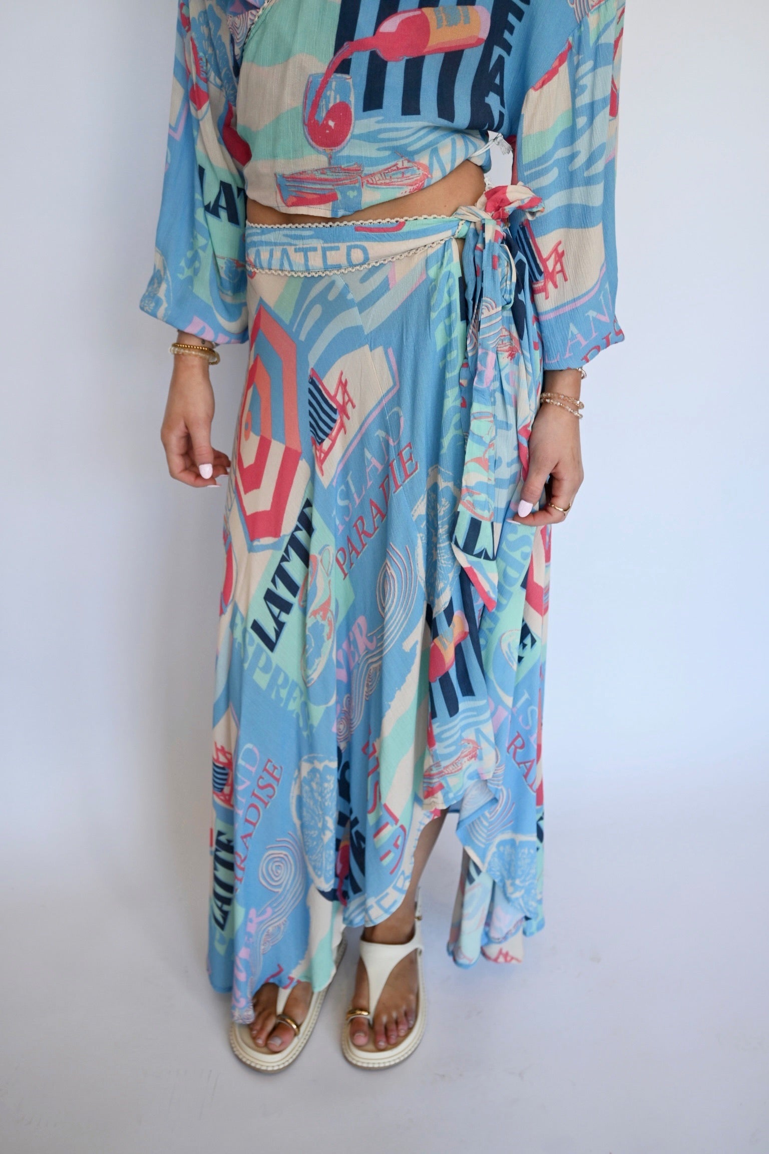 A person wearing a light blue tropical-"blue vacay" themed two-piece set with a matching tie-waist maxi skirt and cropped long-sleeve top, featuring a colorful abstract print with palm trees, flamingos, and text, paired with white slide sandals