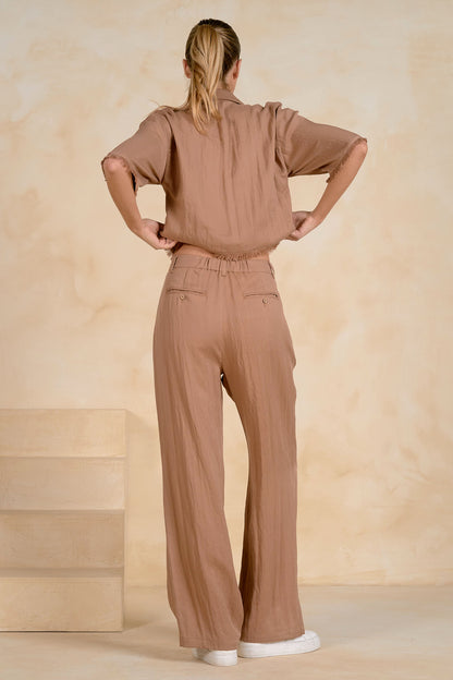 A woman shown from the back wearing a matching taupe-brown short-sleeve fray top and wide-leg trousers set, standing against a textured beige studio backdrop.