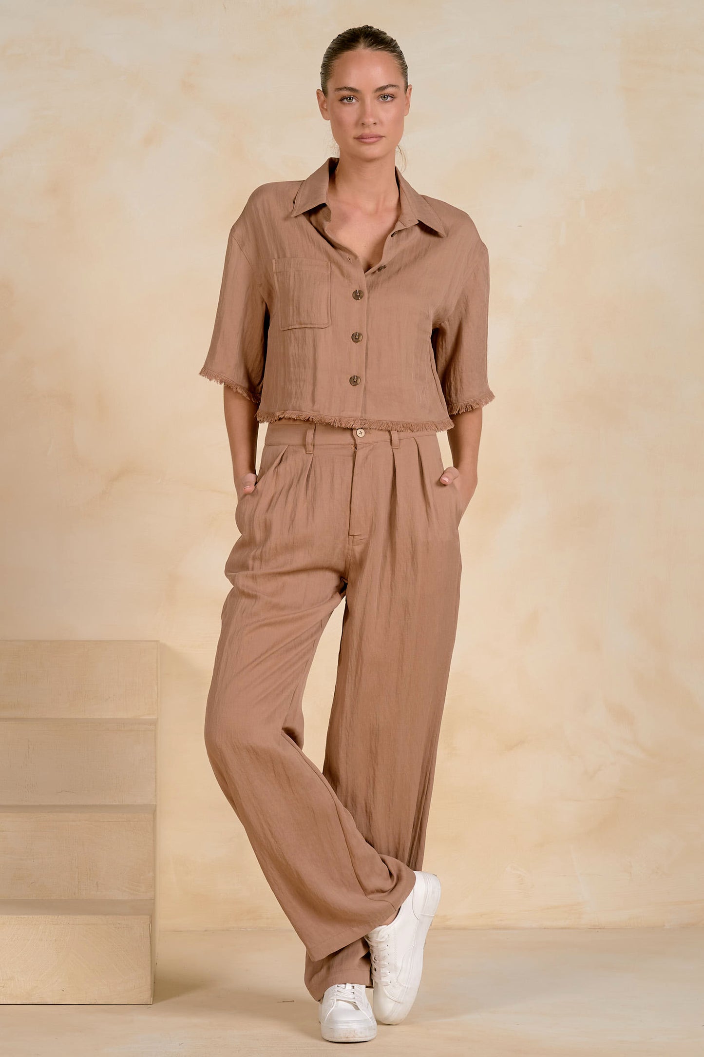 A woman modeling a matching two-piece linen set in a warm taupe-brown color, featuring a short-sleeve button-up fray shirt and wide-leg trousers, styled with white sneakers against a textured beige backdrop.