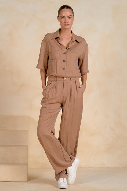 A woman modeling a matching two-piece linen set in a warm taupe-brown color, featuring a short-sleeve button-up fray shirt and wide-leg trousers, styled with white sneakers against a textured beige backdrop.