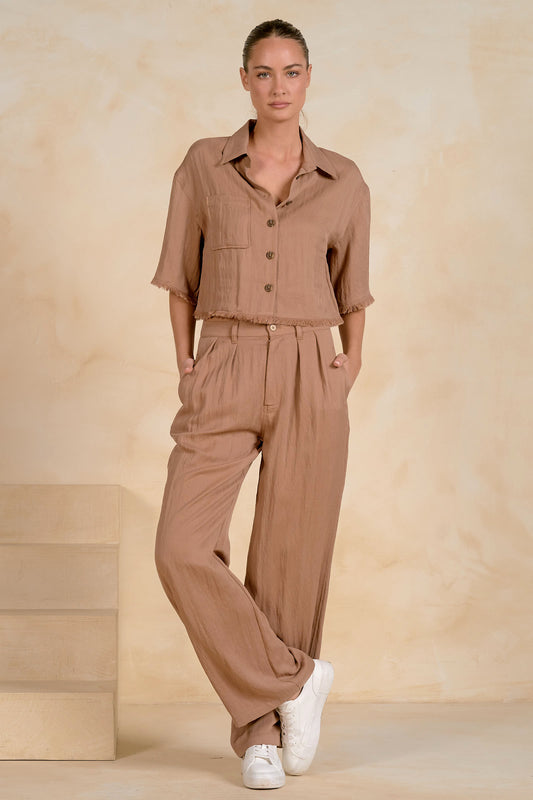 A woman modeling a matching two-piece linen set in a warm taupe-brown color, featuring a short-sleeve button-up fray shirt and wide-leg trousers, styled with white sneakers against a textured beige backdrop.