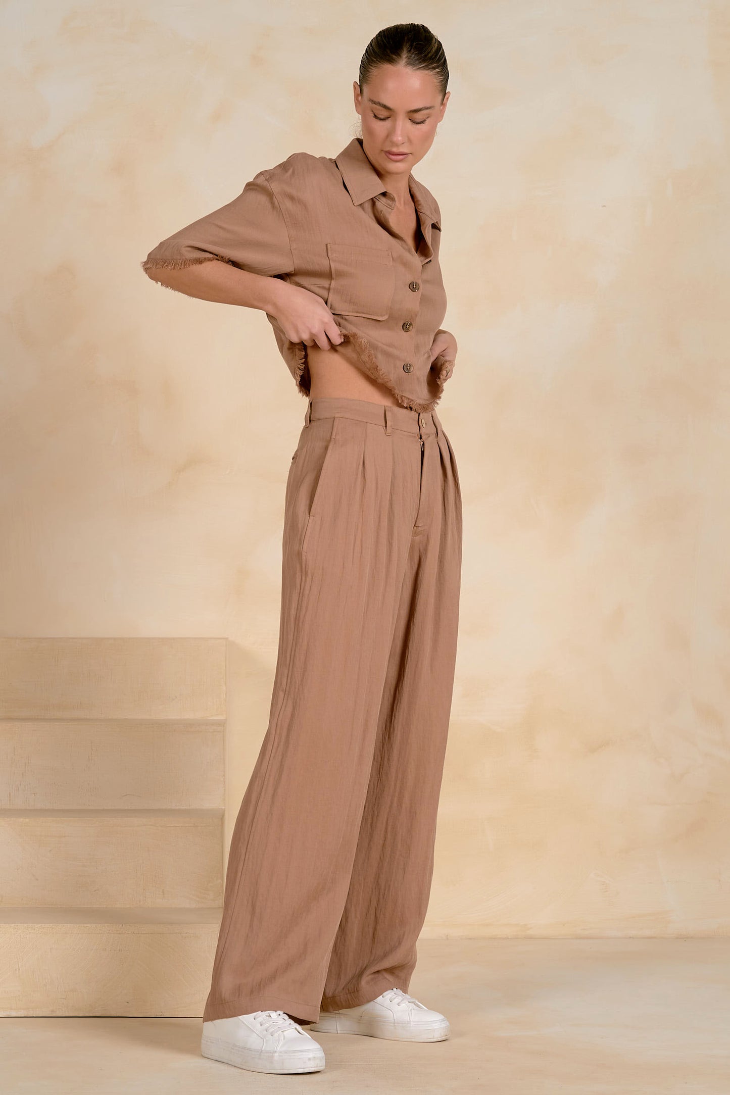 A woman modeling a matching taupe-brown two-piece set consisting of a short-sleeve button-front fray shirt and high-waisted fray wide-leg trousers, styled with white sneakers against a warm beige textured studio backdrop.