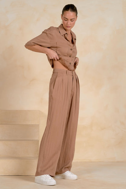 A woman modeling a matching taupe-brown two-piece set consisting of a short-sleeve button-front fray shirt and high-waisted fray wide-leg trousers, styled with white sneakers against a warm beige textured studio backdrop.