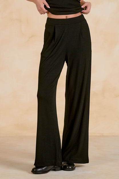 A person wearing black wide-leg pleated pants with a high waistband, paired with black shoes, standing against a warm beige textured studio backdrop.