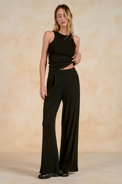 A woman modeling a black sleeveless crop top and matching black wide-leg pants against a warm beige textured studio backdrop.