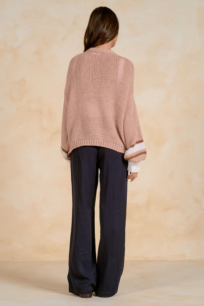 A woman shown from the back wearing a tan  knit sweater with white striped cuffs and dark navy wide-leg trousers, standing against a textured beige studio backdrop.