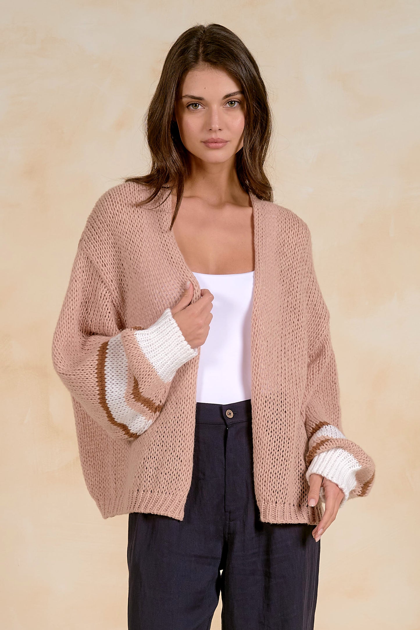 A woman modeling a chunky knit cardigan in a tan color, featuring contrasting white ribbed striped cuffs and a white inner layer, paired with dark trousers against a textured beige background.