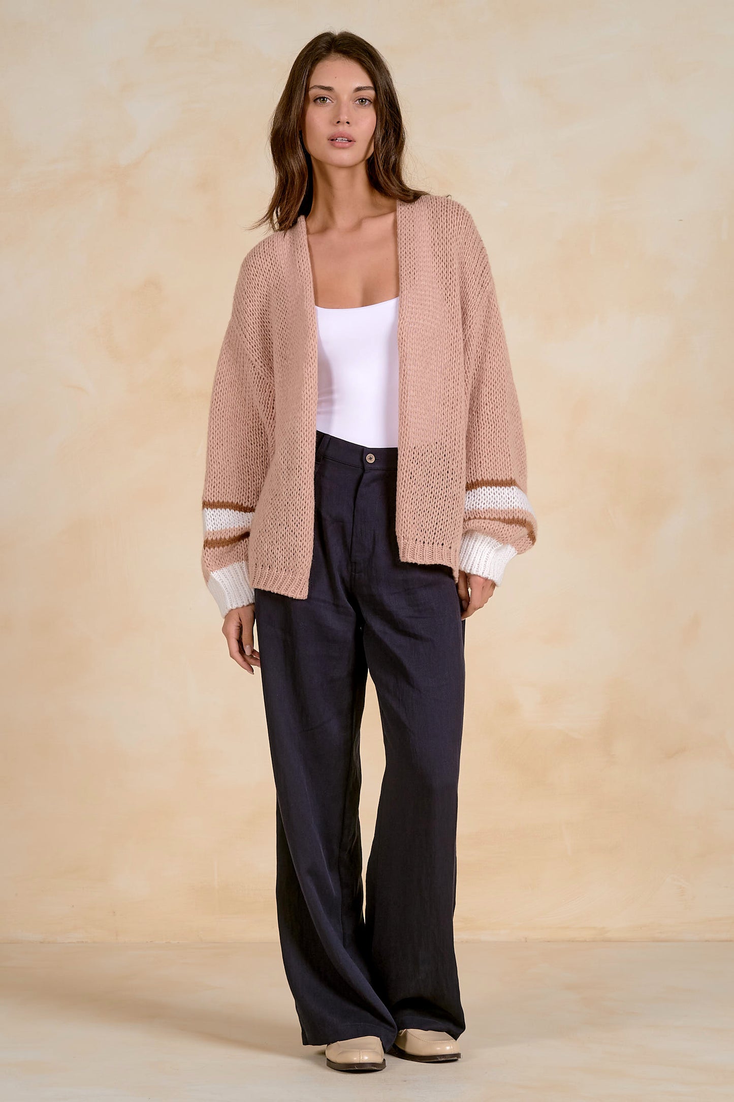 A woman stands against a warm beige textured backdrop wearing a tan open-front knit cardigan with cream striped cuffs, a white tank top, dark navy wide-leg trousers, and beige flats.
