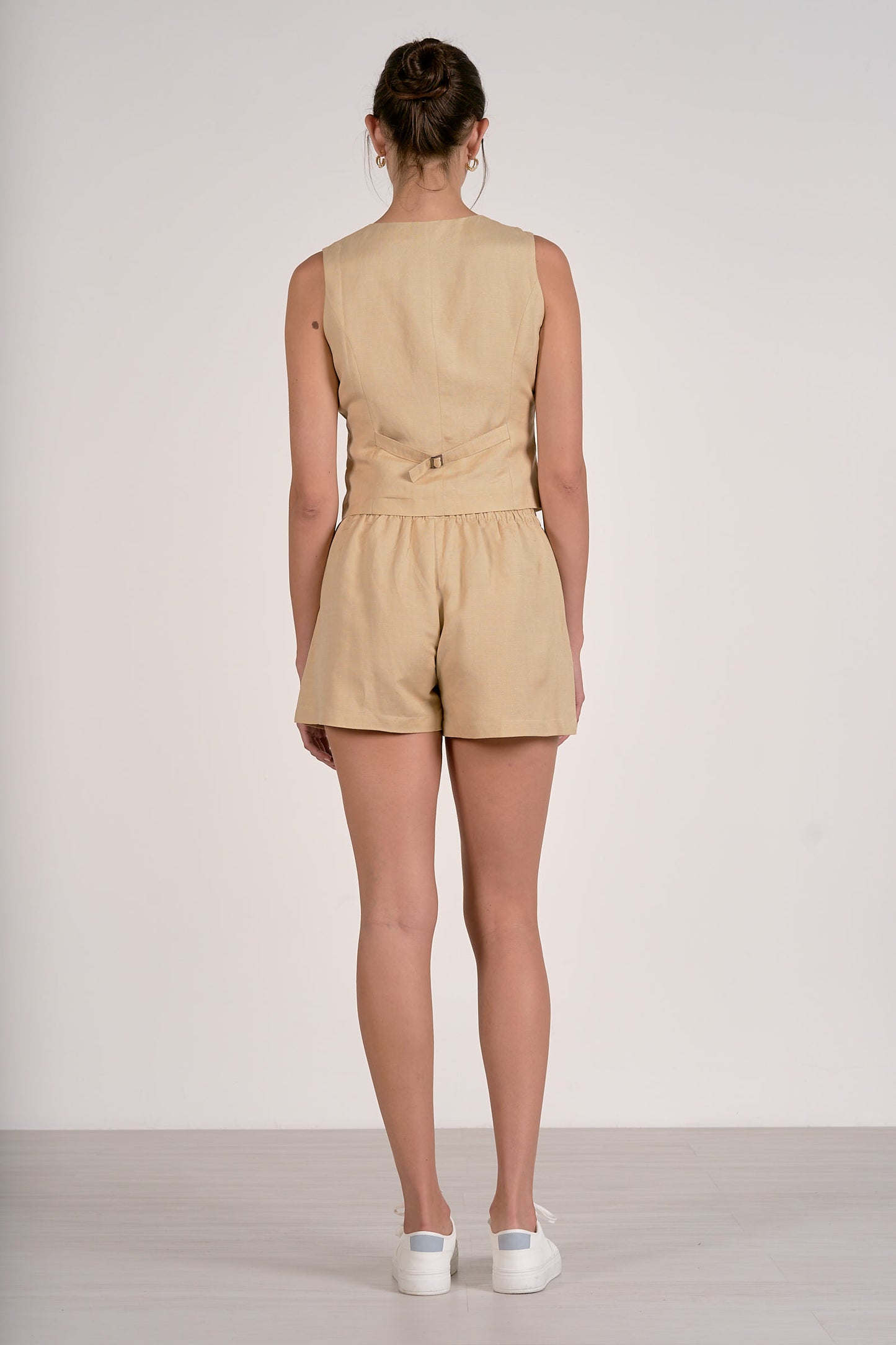 Back view of woman wearing an oatmeal sleeveless vest and shorts on a plain background