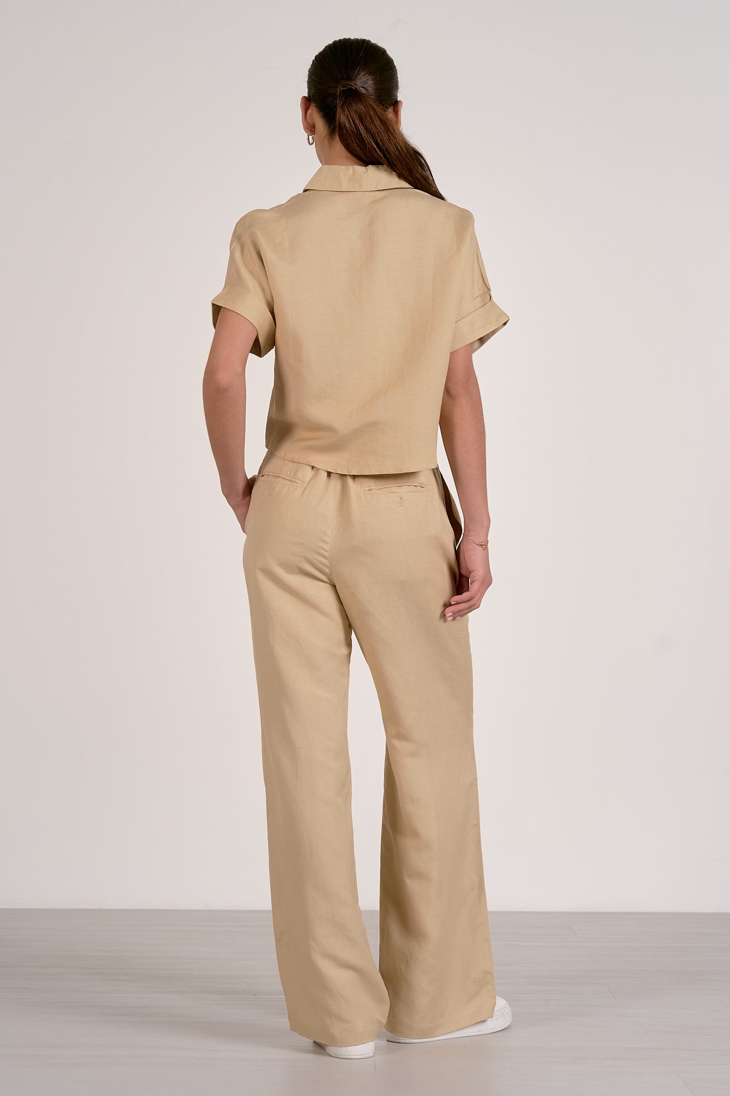 Back view of woman wearing an oatmeal colored outfit with a button up, collared shirt and oatmeal pleated pants on a plain background