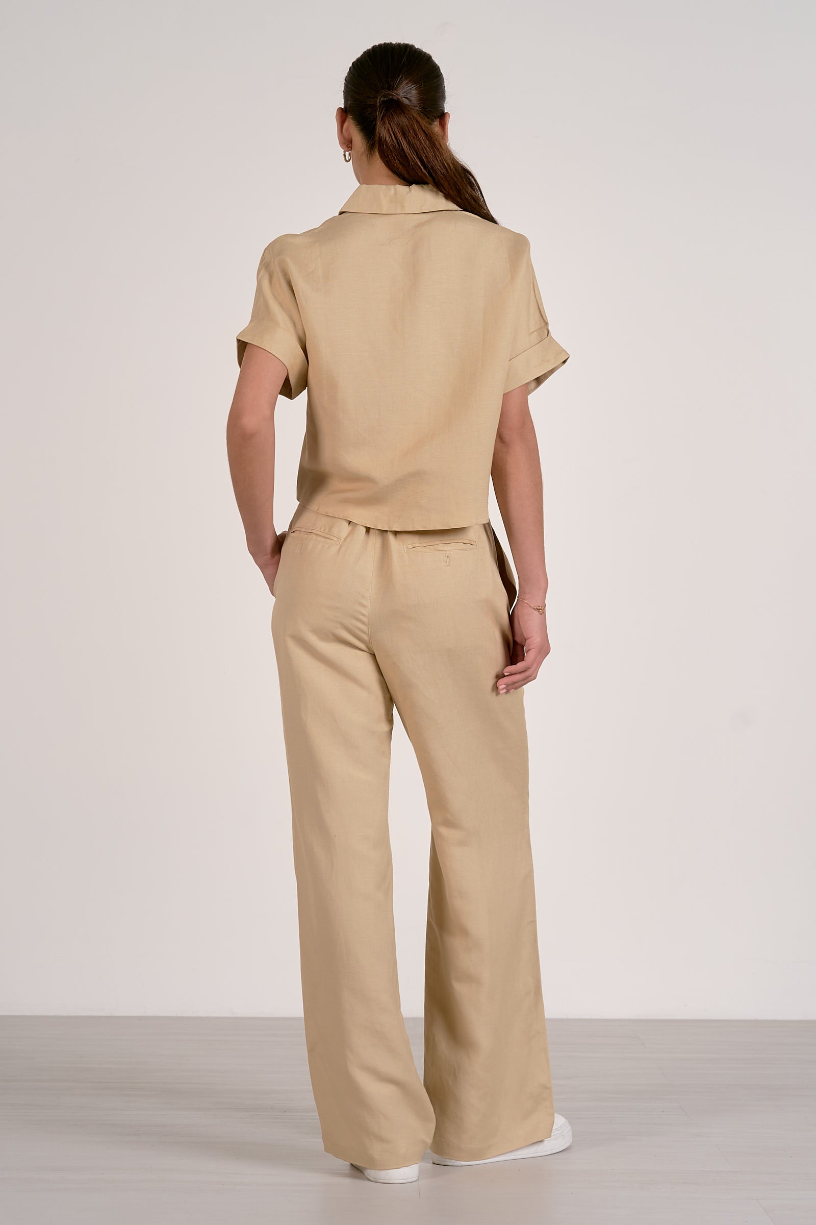 Back view of woman wearing an oatmeal colored outfit with a button up, collared shirt and oatmeal pleated pants on a plain background