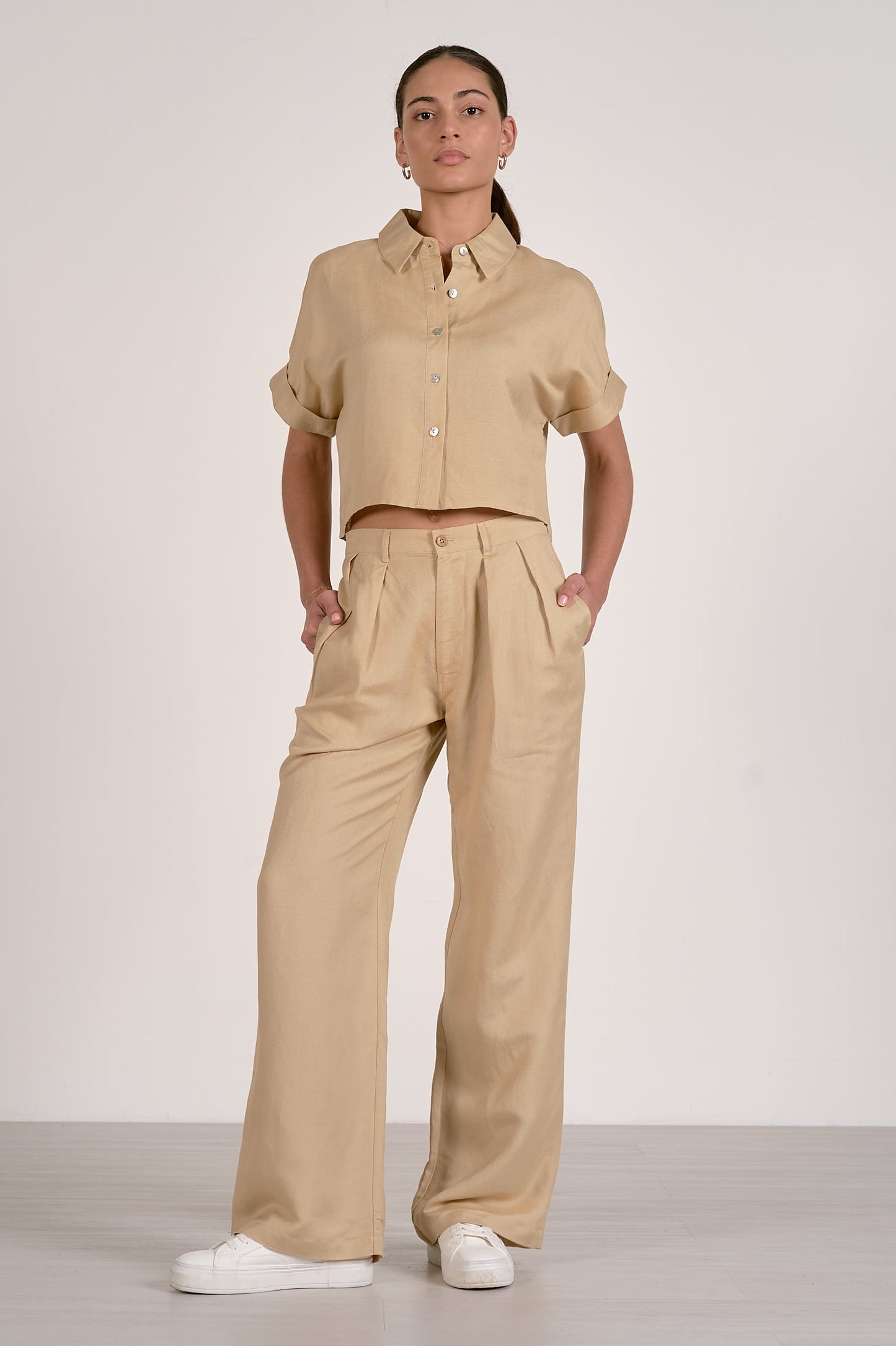 Front view of woman wearing an oatmeal colored outfit with a button up shirt and oatmeal pleated pants on a plain background