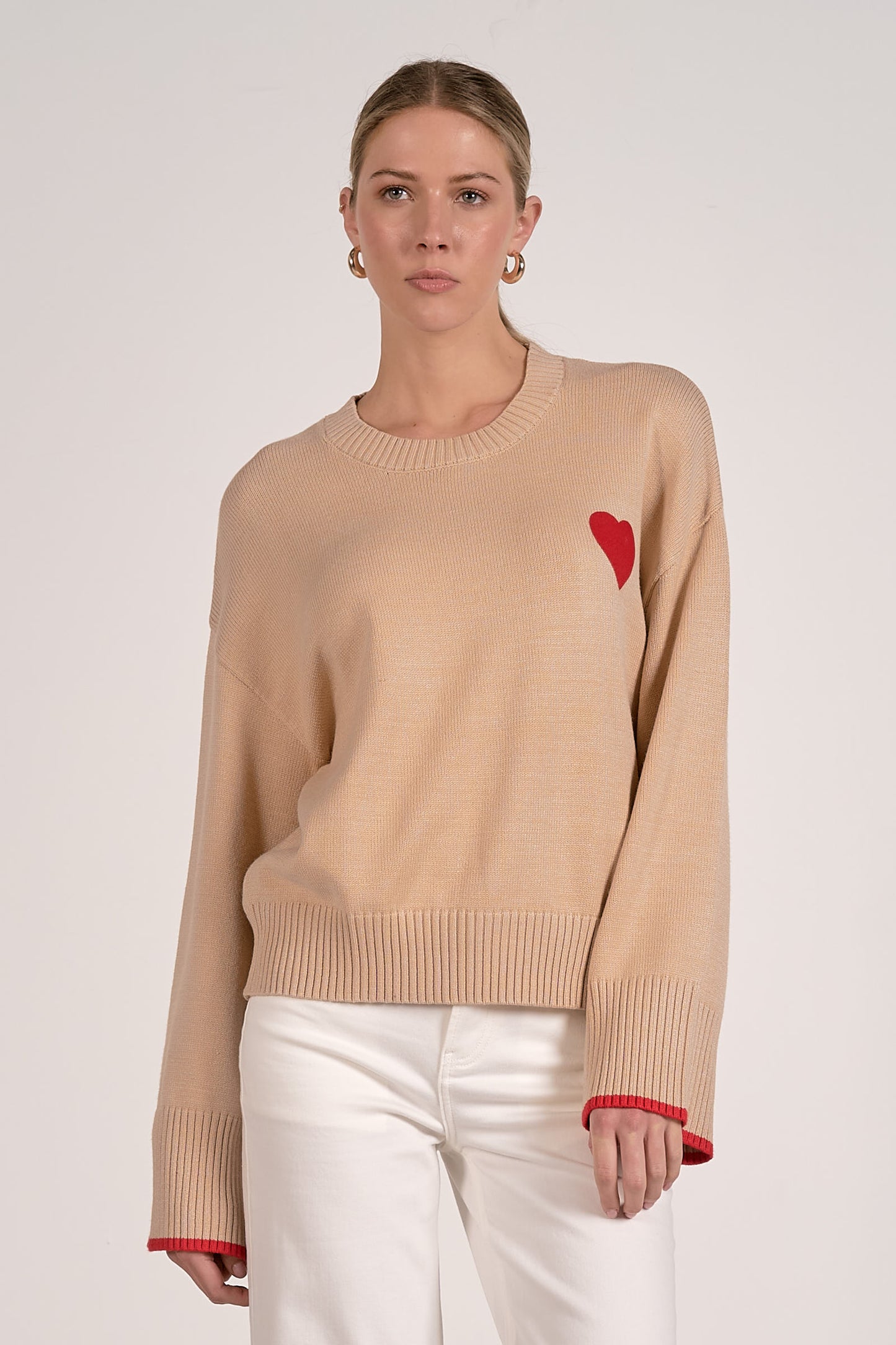 A woman modeling a tan crewneck sweater with a small red heart on the left chest and red trim at the cuffs, paired with white pants, against a light gray studio background.